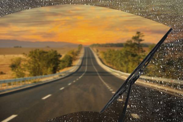 A first-person view of a driver on a long road behind a windshield with wipers clearing away the rain.