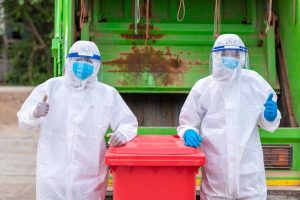 Two people in personal protective equipment, gloves, and face masks give the thumbs up while standing by a blue trash bin.