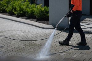 A worker using a pressure washing jet to spray water onto a paver walkway outside a business lined with bushes.