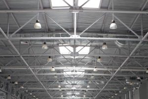 A modern gray warehouse ceiling, with a row of skylights down the center and pendant lights hanging evenly all across.