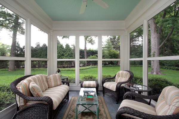 An enclosed porch with three walls of screen panels and black wicker furniture facing a backyard with several tall trees.
