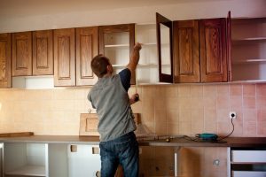 A man is doing some work in the kitchen. He's measuring the length of the cabinet to replace cabinet doors.
