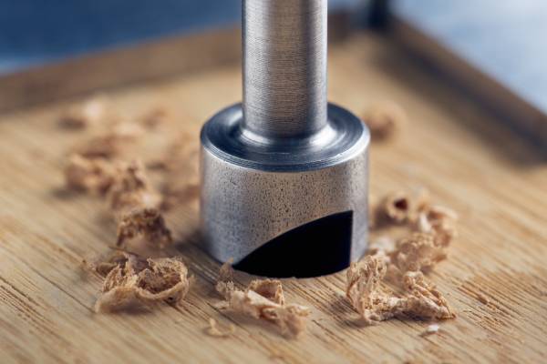 A countersink drill bit with a steel triangle ruler attachment makes a hole inside an oak wood plank.
