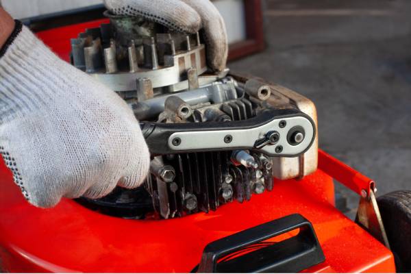 A cropped image of hands wearing white gloves turning a socket wrench on a red lawn mower engine for repairs.