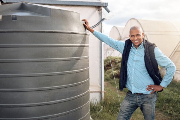 A farmer wearing a vest and jeans stands next to a large water tank on his property with a smile on his face.