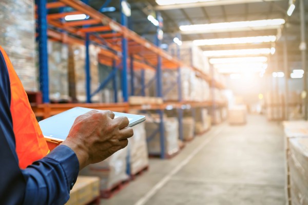 A person stands slightly out of view, wearing a blue shirt and orange safety vest. They're holding a tablet in a warehouse.