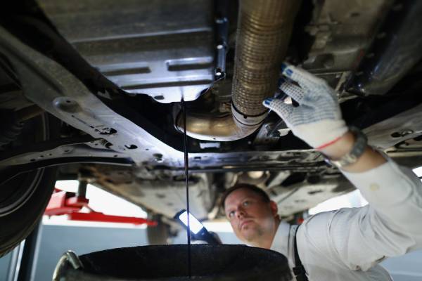 An automotive technician wearing gloves holding a flashlight beneath a vehicle’s undercarriage performing an oil change.