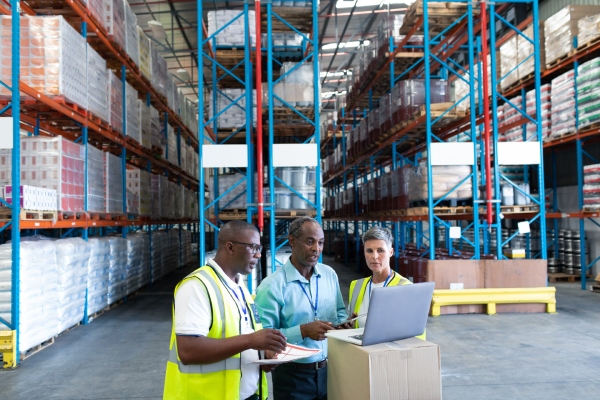 Three staff members stand in a warehouse and look at a laptop together as they discuss operating costs.
