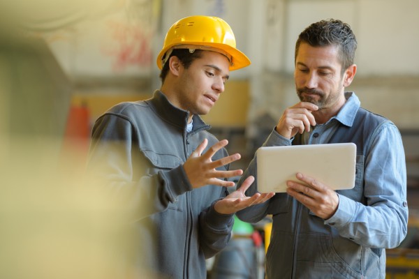 A worker holds up a tablet, looking at some work information with another worker in a yellow hard hat.