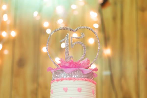 A white cake with pink hearts frosted around it has a sparkly heart standing on top of it with a number 15 at the center.