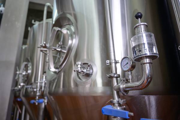 Large stainless steel brewing tank with a pressure valve, tubes, and safety locks next to other brewing tanks.