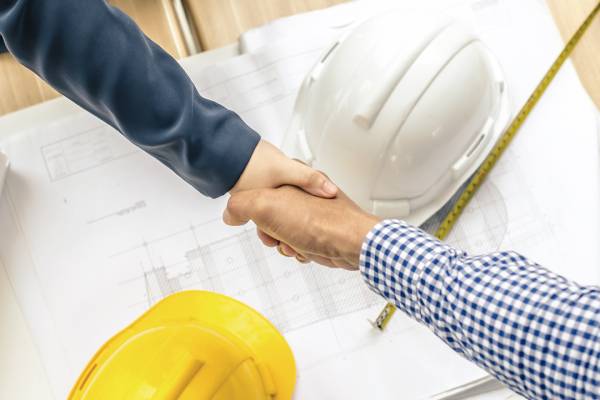 Two hands shaking over a blueprint sitting next to a pair of white and yellow hard hats and a tape measure on table.