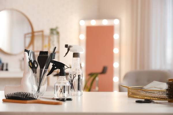 Hairdressing tools, including a brush, a spray bottle, and scissors, on a desk with a standing Hollywood vanity mirror in the background.