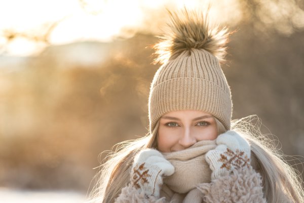 A woman standing outside in a cozy jacket and winter beanie. She's pulling her knitted scarf up over her face.