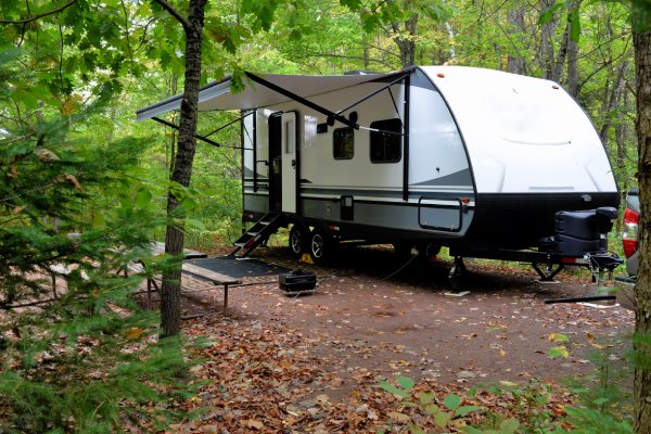 A white travel camper is parked in the woods with a protective visor extended from its side. No people are present.