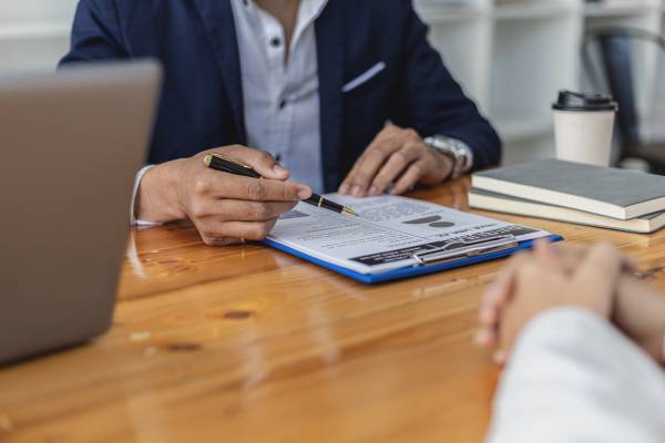An interviewer at a desk gesturing to a resume on a clipboard while speaking to an applicant with his hands on the desk.