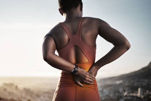 A woman wearing athletic clothing touches the back of her lower right hip with both her hands while she is outdoors.