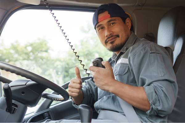 A smiling truck driver wearing a backward baseball cap shows a thumbs up with a wired radio in his other hand.