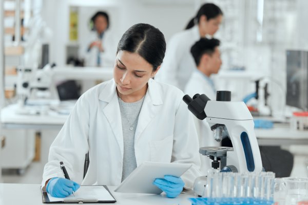 A female scientist wearing a white robe and gloves writing in a notebook beside a microscope and sample tubes.