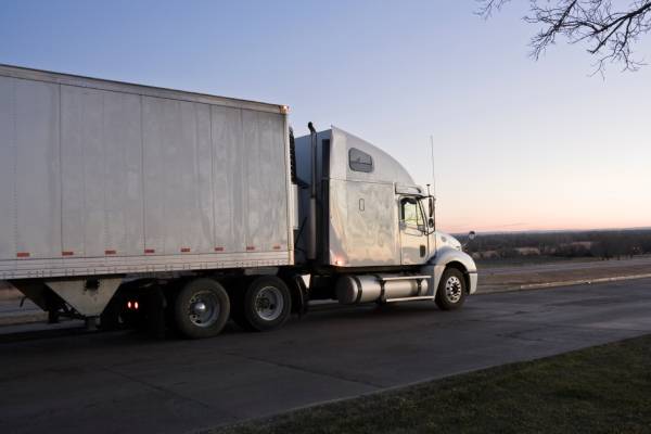 A white semi-truck drives down the empty road at sunrise with a little bit of pink in the blue sky.