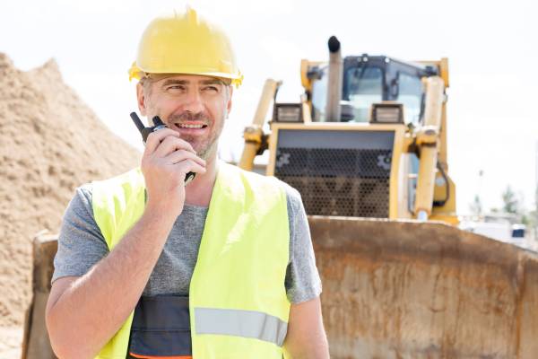 A construction site supervisor in a hard hat and a vest. He's using a two-way radio to communicate with equipment behind him.
