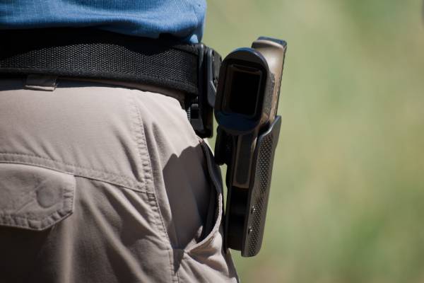 A person wearing khakis and a blue shirt standing with a belt attached to a holster and gun in front of a blurry background.