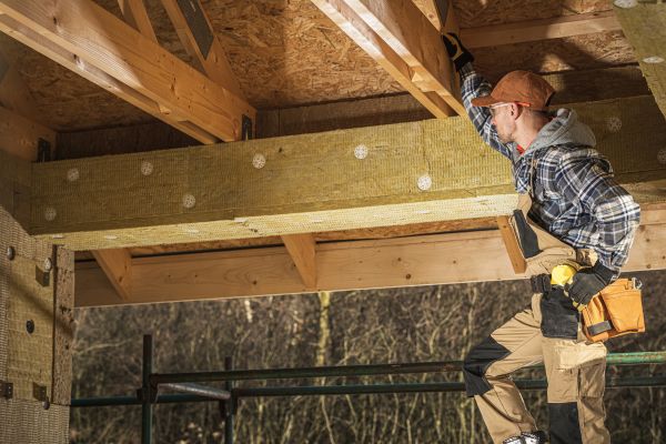 A worker in a plaid shirt, beige overalls, and gloves standing on scaffolding, reaching toward a wooden roof frame.