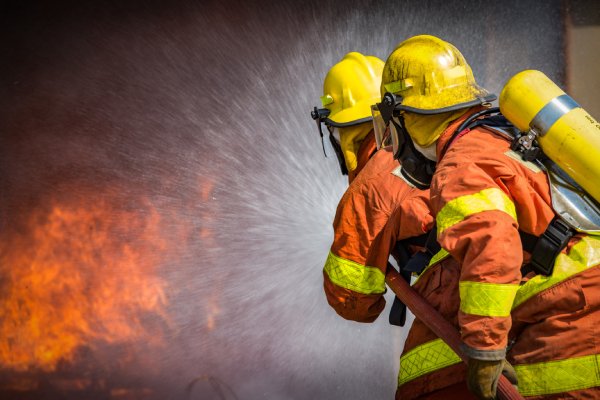 Two firefighters in orange and yellow uniforms and yellow hard hats spray a hose toward smoky flames.