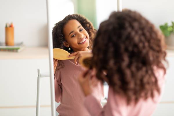 A young girl with textured hair smiling as she stands in front of a full-length mirror and carefully brushes her curls.