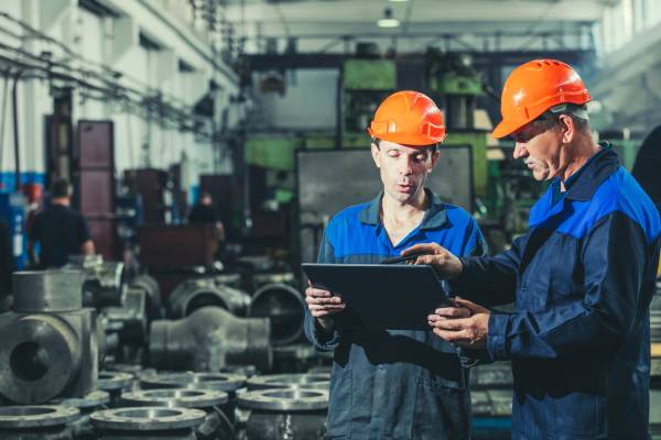 Two workers in a factory wearing hardhats and body suits holding an electronic tablet and pointing to the display.