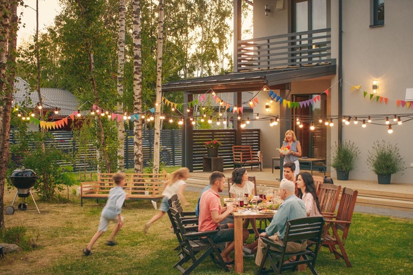 At a backyard party, a group of people sit at a table, talking and eating. Two children run around the table.