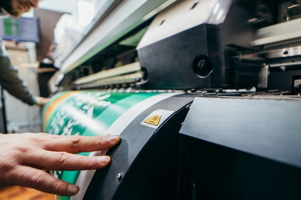 A printing technician holds the sides of a large format graphic print project as it exits the machine.