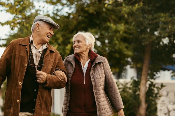 A senior man and woman link arms and walk together. They both wear jackets and look at each other while smiling.