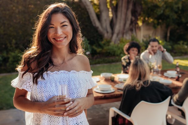A woman stands outside, holds a glass, and smiles toward the camera. She stands in front of a table with guests.