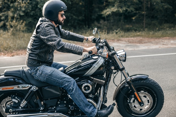 A close-up of a man wearing a black helmet, a leather jacket, and blue jeans while riding a motorcycle on a paved road.