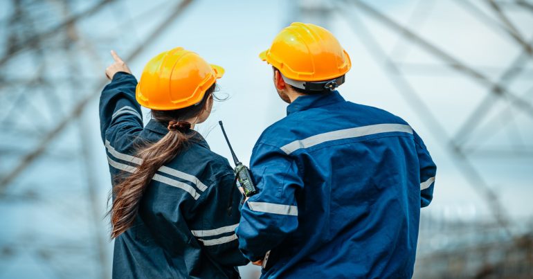 Two industrial workers wearing blue clothing and yellow hard hats face an electrical tower. One of them points at it.