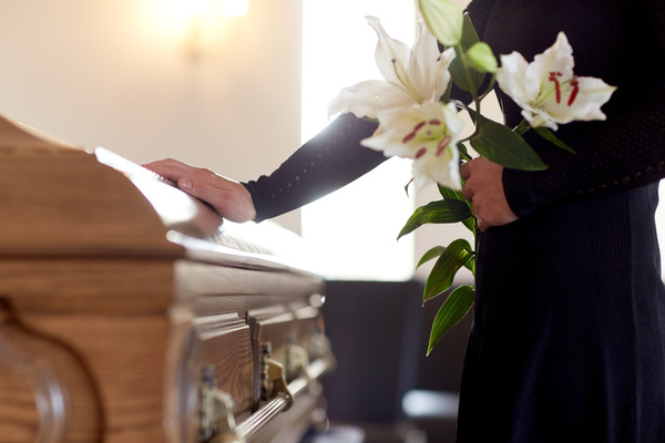 A person at a funeral holds flowers in their left hand and rests their right hand on the closed coffin in front of them.