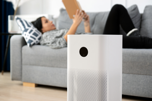 A close up of an air purifier in the living room. A female homeowner is laying on the couch behind it reading a book.