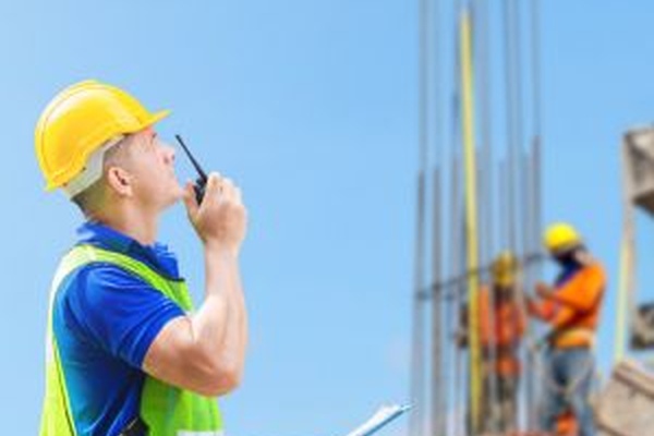 A man in a yellow hard hat and green safety vest uses a radio and looks up. Behind him are workers building structural supports.
