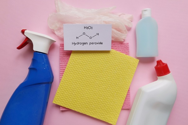 A card reading "Hydrogen peroxide" rests on latex gloves and two cleaning pads, surrounded by three cleaning product bottles.