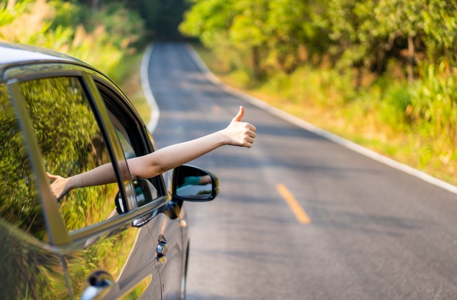 A close-up showing someone giving a thumbs-up out of the window of a small black car driving down a tree-lined road.