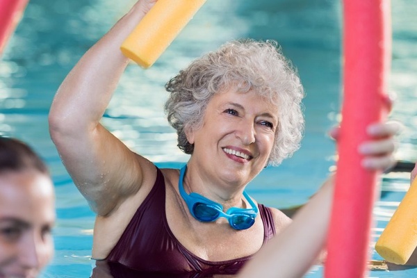 A grinning senior woman in a swimsuit holds a yellow pool noodle over her head and leans to the side in a water aerobics class.