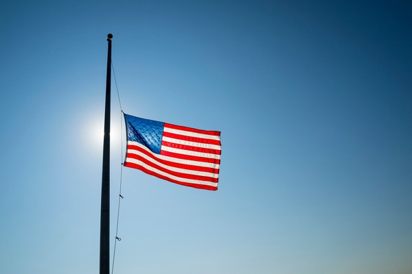 An American flag flies in the wind at half staff on a flagpole. The sun shines through the flag in the background.