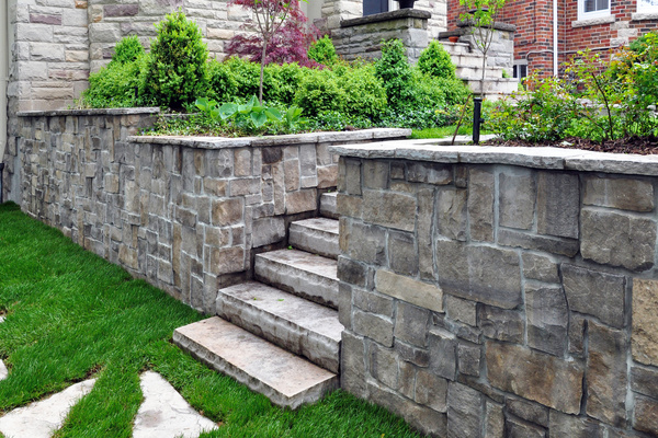 A concrete retaining wall and stairs made out of stones leading to a backyard filled with green plants.