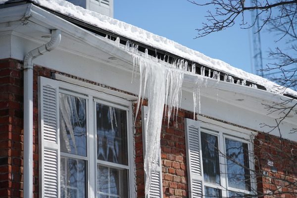 Large icicles hang from the gutters and windows of a house in winter. The gutter is bending from the ice.