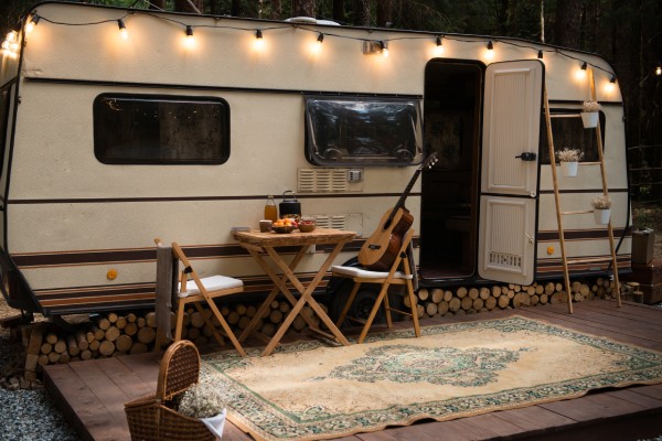 A long white camper with string lights hanging from it. A small table and chairs with a guitar are set up by the front door.