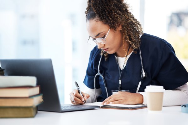 A woman in scrubs with a stethoscope around her neck sits in front of a laptop and writes with a pen in a journal.