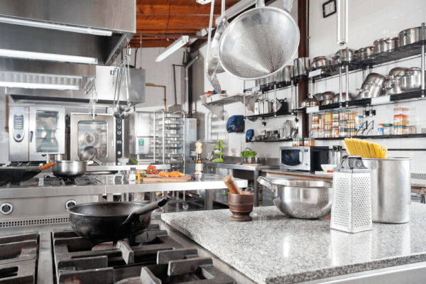 A commercial kitchen with utensils on the counter, a pan on the stove, and a shelf of pots and pans on the wall.