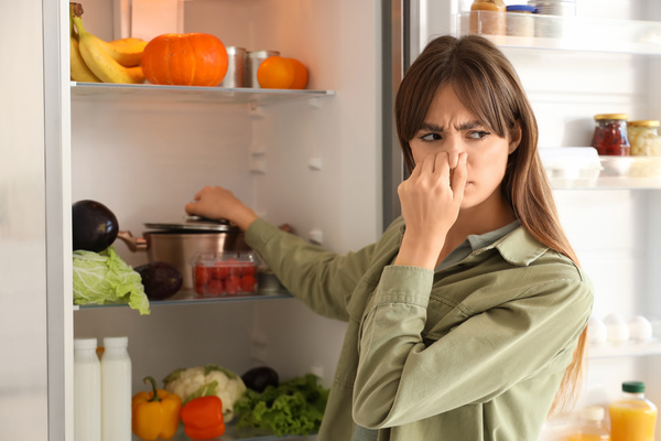 A woman pinches her nose with her hand and furrows her eyebrows while standing in front of an open refrigerator.
