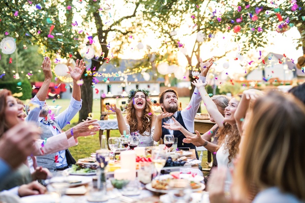 A smiling bride and groom sitting at an outdoor table and catching confetti with their laughing guests.